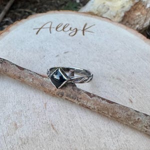 May include: A silver ring with a black, diamond-shaped stone. The ring has a detailed, vine-like design on the band. The ring is displayed on a wooden stick, with the name "Allyk" written on the wooden surface in the background.