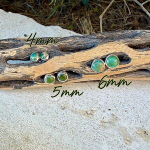 May include: Round stud earrings featuring green turquoise stones set in silver bezels. The earrings are displayed in three sizes: 4mm, 5mm, and 6mm, with the size labels clearly written on the wood surface. Each stone displays unique natural veining.