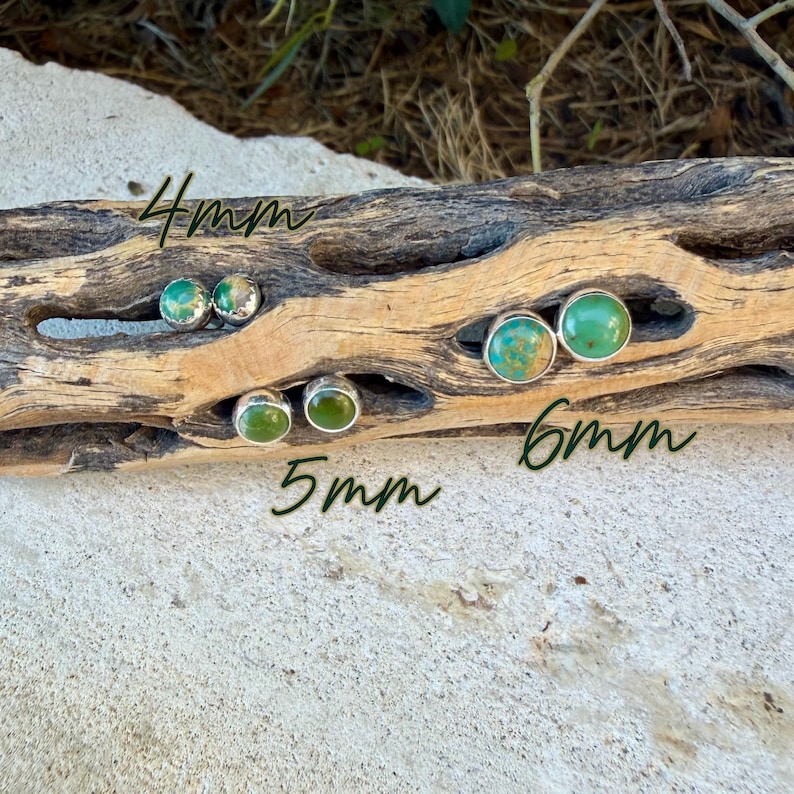 May include: Round stud earrings featuring green turquoise stones set in silver bezels. The earrings are displayed in three sizes: 4mm, 5mm, and 6mm, with the size labels clearly written on the wood surface. Each stone displays unique natural veining.