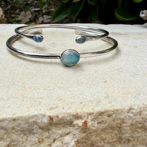 K&ouml;nnte beinhalten: Zwei silberfarbene Armreifen mit ovalen, irisierenden Edelsteinen. Ein Armreif hat einen gr&ouml;&szlig;eren Edelstein in der Mitte, w&auml;hrend der andere zwei kleinere Edelsteine hat. Der Schmuck wird auf einer hellen, strukturierten Oberfl&auml;che pr&auml;sentiert.