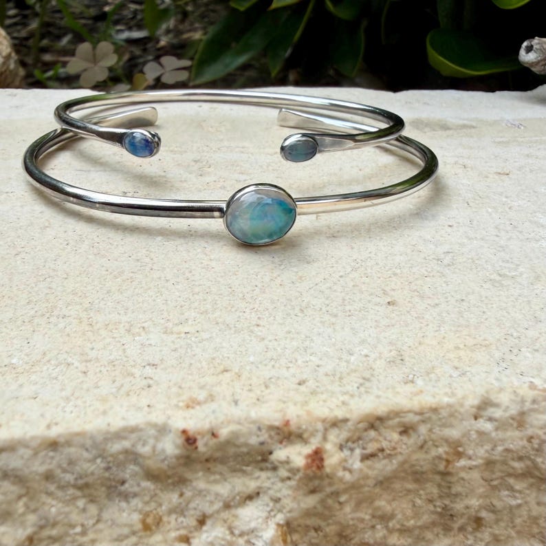 K&ouml;nnte beinhalten: Zwei silberfarbene Armreifen mit ovalen, irisierenden Edelsteinen. Ein Armreif hat einen gr&ouml;&szlig;eren Edelstein in der Mitte, w&auml;hrend der andere zwei kleinere Edelsteine hat. Der Schmuck wird auf einer hellen, strukturierten Oberfl&auml;che pr&auml;sentiert.