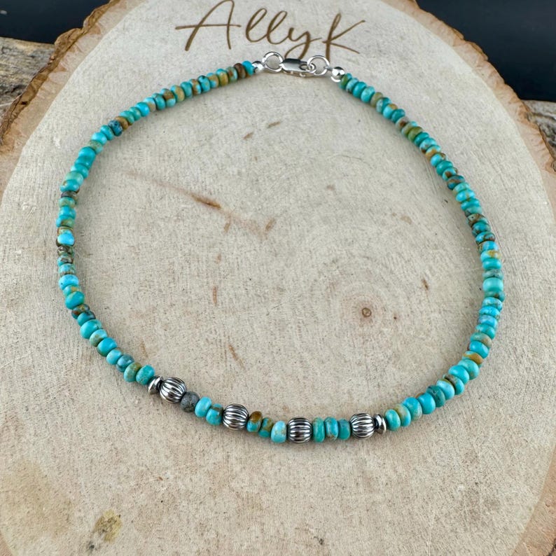 May include: Turquoise beaded anklet with silver accents. The anklet features small, round turquoise beads strung together, interspersed with silver-colored, ridged beads. The clasp is silver. The anklet is displayed on a wooden surface with the name "Allyk" written on it.