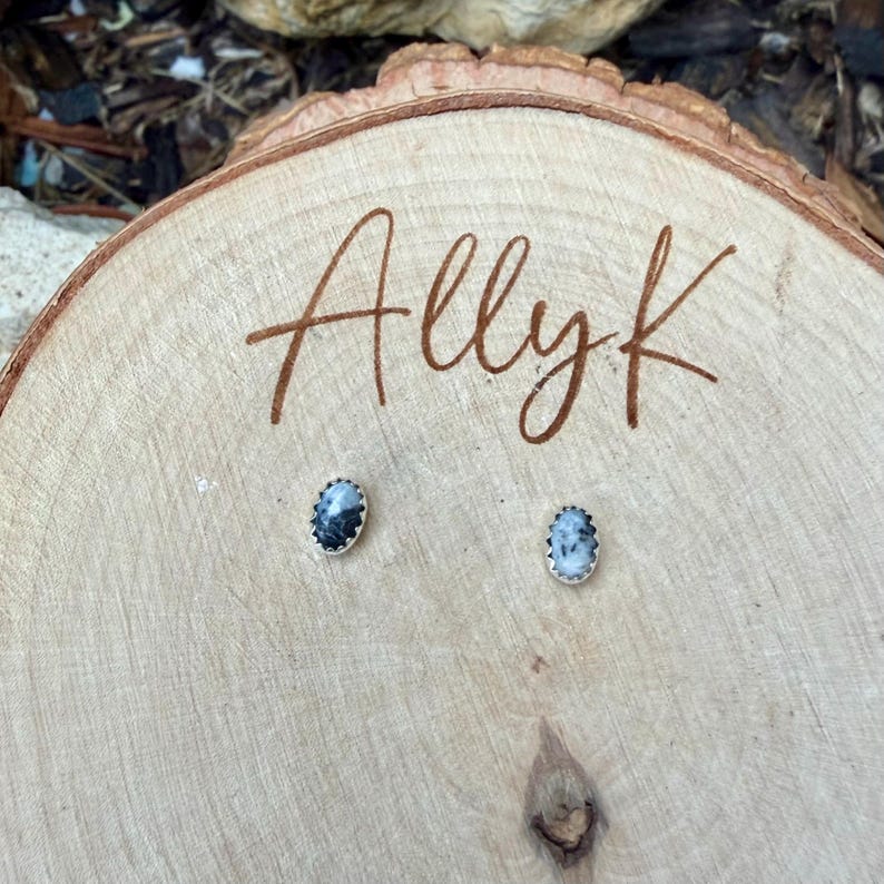 May include: A pair of oval-shaped stud earrings with a black and white stone set in a silver-colored metal. The earrings are displayed on a wooden surface with the handwritten text "Allyk". The wood has a natural grain and a light brown color.