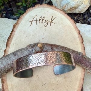 Puede incluir: Una pulsera de pu&ntilde;o de cobre con un dise&ntilde;o floral grabado. La pulsera se muestra sobre una rodaja de madera con el nombre "Allyk" escrito. La pulsera mide aproximadamente 3,8 cm de ancho.