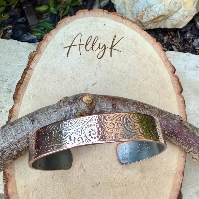 Puede incluir: Una pulsera de pu&ntilde;o de cobre con un dise&ntilde;o floral grabado. La pulsera se muestra sobre una rodaja de madera con el nombre "Allyk" escrito. La pulsera mide aproximadamente 3,8 cm de ancho.