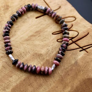 May include: A beaded bracelet featuring pink and black gemstone beads. The bracelet has silver-colored metal accents and is strung on a flexible cord. The beads are oval-shaped and vary in color, creating a unique pattern. The bracelet is displayed on a wooden surface.