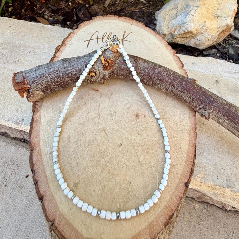 May include: A delicate white beaded necklace with silver accents. The necklace is displayed on a wooden surface with the handwritten text "Alle K". The beads are small and evenly spaced, creating a simple yet elegant design.