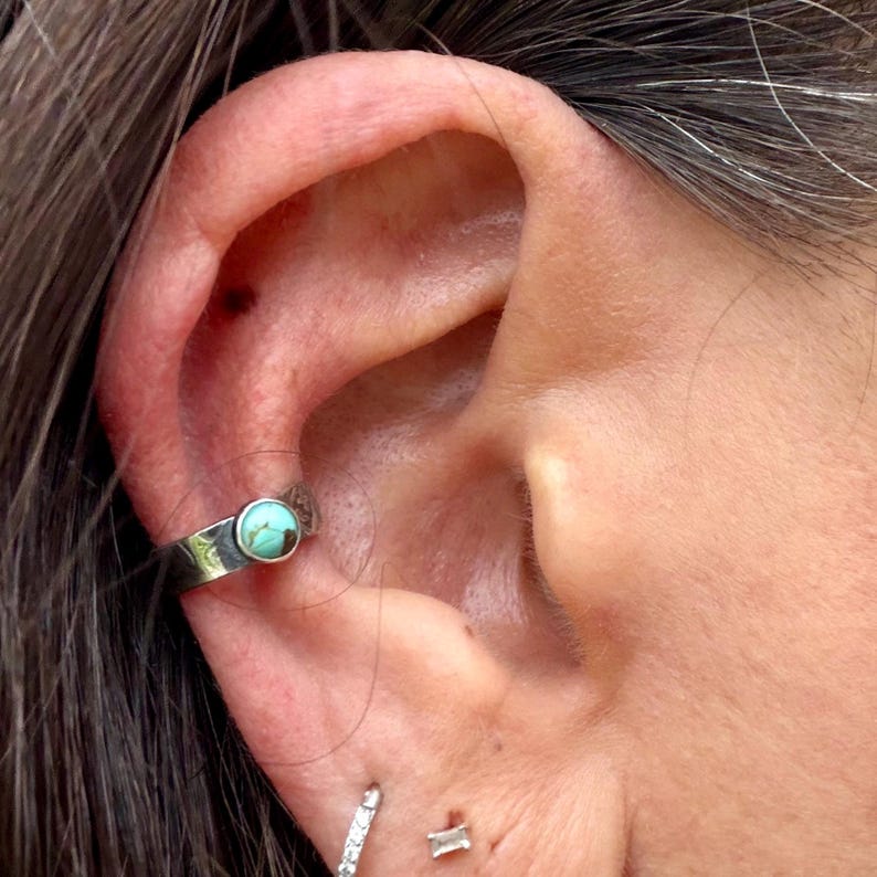 K&ouml;nnte beinhalten: Ein silberner Ohrklemmen-Schmuck mit einem runden, t&uuml;rkisfarbenen Stein. Der Schmuck wird am oberen Ohrknorpel getragen. Weitere Ohrringe sind im Ohrl&auml;ppchen sichtbar.