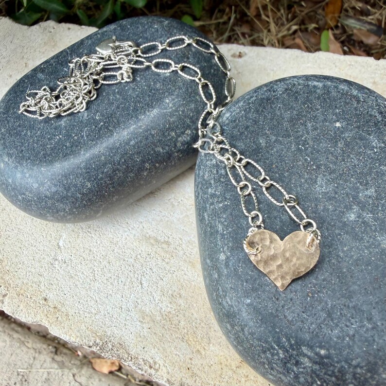 May include: A silver-toned necklace featuring a hammered heart-shaped pendant suspended from a textured oval link chain. The metal heart has a dimpled, uneven surface texture and is attached to the chain by two small jump rings on each side.