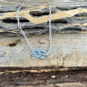May include: A silver necklace featuring a rectangular pendant with a cutout of the state of Texas. The pendant is connected to a chain necklace. The background is a piece of weathered wood.