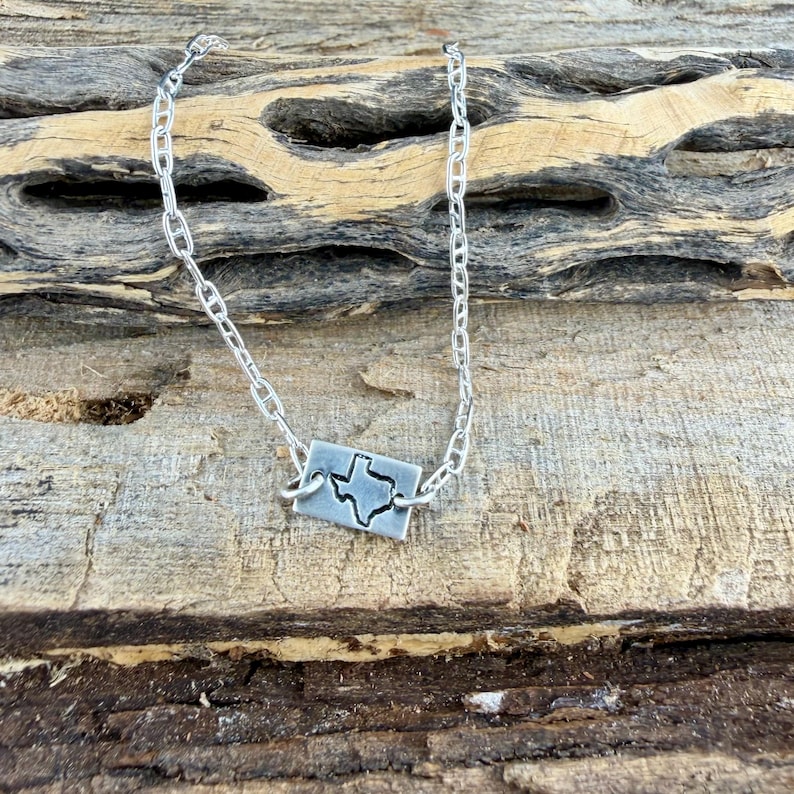 May include: A silver necklace featuring a rectangular pendant with a cutout of the state of Texas. The pendant is connected to a chain necklace. The background is a piece of weathered wood.