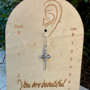 May include: A silver-toned metal earring shaped like a dagger with an ornate cross-guard, suspended from a hook. The earring is displayed on a light-coloured wooden card featuring an ear outline, cm markings, and the text You are beautiful.