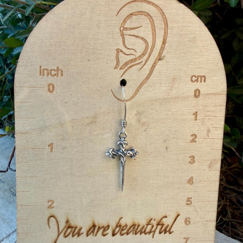 May include: A silver-toned metal earring shaped like a dagger with an ornate cross-guard, suspended from a hook. The earring is displayed on a light-coloured wooden card featuring an ear outline, cm markings, and the text You are beautiful.