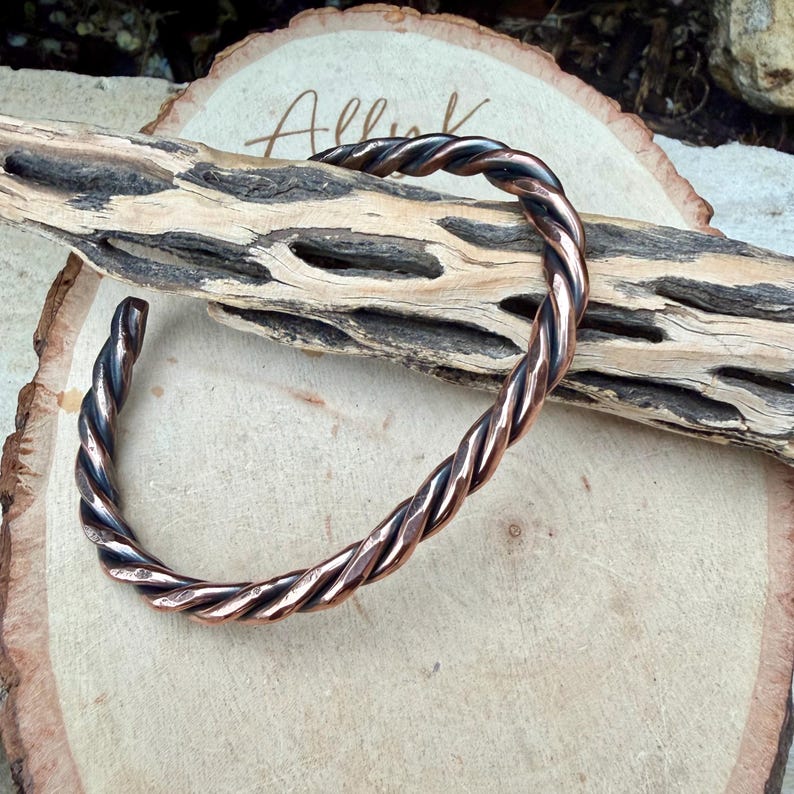 May include: A copper-colored cuff bracelet with a twisted rope design. The open-ended bracelet is displayed on a wooden surface with a natural, organic aesthetic. The metal has a polished, reflective quality.