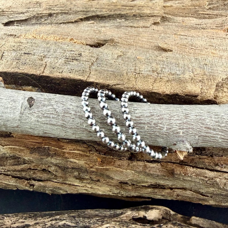 Puede incluir: Tres anillos de cuentas plateadas se exhiben sobre un trozo de madera flotante. Los anillos est&aacute;n hechos de peque&ntilde;as cuentas redondas ensartadas para formar un c&iacute;rculo continuo. La madera flotante tiene un color gris claro y una textura rugosa.