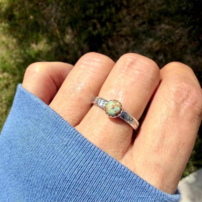 May include: A silver ring with a round, light turquoise stone set in the center. The band has a textured design and is worn on a finger. The ring is a piece of jewelry.