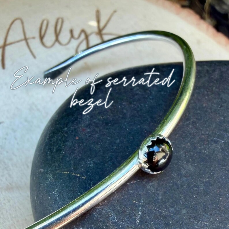 Op de afbeelding: Een zilverkleurige armband met een zwarte edelsteen in een gekartelde bezel. De gebogen armband heeft de tekst "Example of serrated bezel" in witte letters.