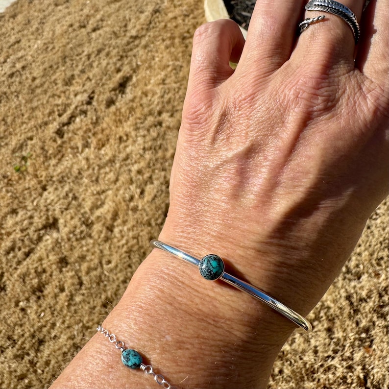 May include: A silver bracelet with a single turquoise stone. A matching silver chain bracelet with turquoise stones is also visible. A silver ring with a feather design is on the hand.