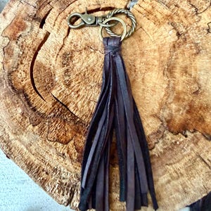 May include: Dark brown leather tassel keychain with a bronze-colored clasp and ring. The tassel is made of long, thin strips of leather, knotted at the top. The keychain is designed for attaching to keys or bags.