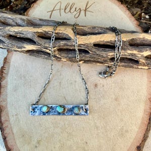 May include: A silver necklace with a rectangular pendant featuring three turquoise stones. The pendant has a textured surface and is suspended from a delicate chain. The necklace is displayed on a wooden surface, with the handwritten text "AllyK" visible.
