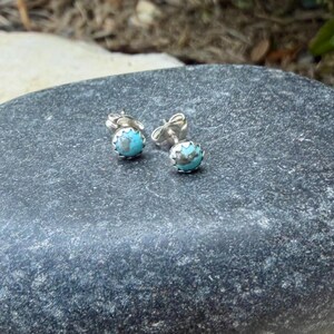 May include: A pair of small, round stud earrings. Each earring features a light blue stone with a silver-colored setting. The earrings are displayed on a dark gray, textured surface.