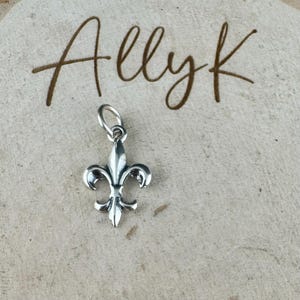 May include: A silver-colored fleur-de-lis charm with a loop for attaching to a necklace or bracelet. The charm is displayed on a light-colored, textured surface with the handwritten text "Allyk" in brown.