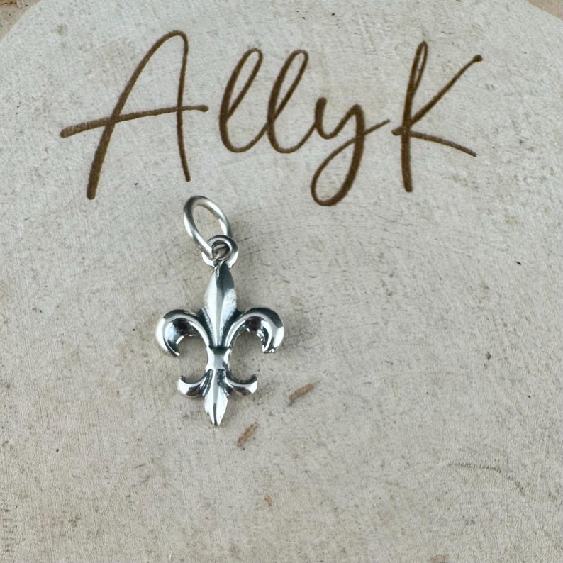 May include: A silver-colored fleur-de-lis charm with a loop for attaching to a necklace or bracelet. The charm is displayed on a light-colored, textured surface with the handwritten text "Allyk" in brown.