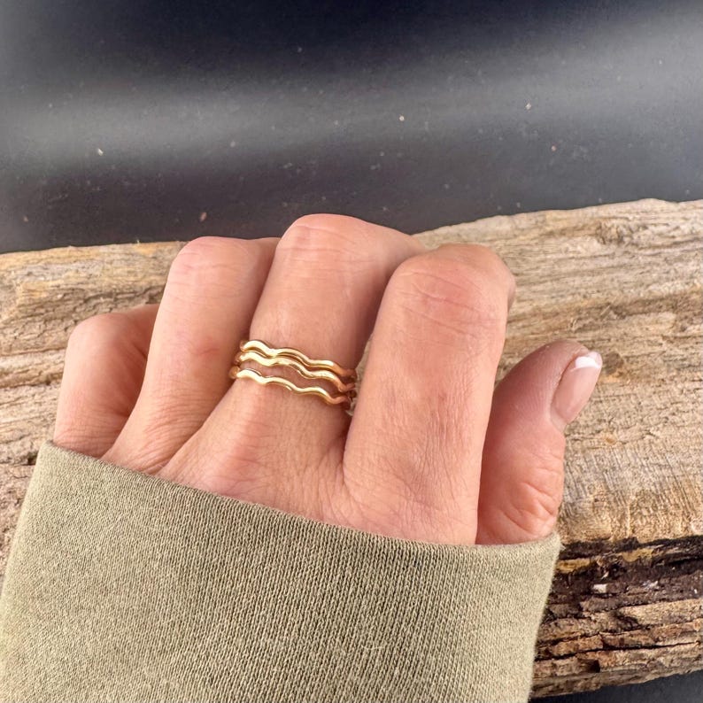 May include: A gold-colored ring with a wavy design. The ring has three bands that create a layered effect. The ring is on a person's finger, and the background is a piece of wood.