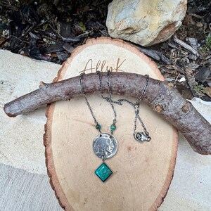 Op de afbeelding: Een zilveren ketting met een munt hanger en een turkoois edelsteen. De ketting heeft een ketting met een sluiting en wordt gepresenteerd op een houten oppervlak met de tekst "Allyk". De turkoois edelsteen heeft een vierkante vorm.