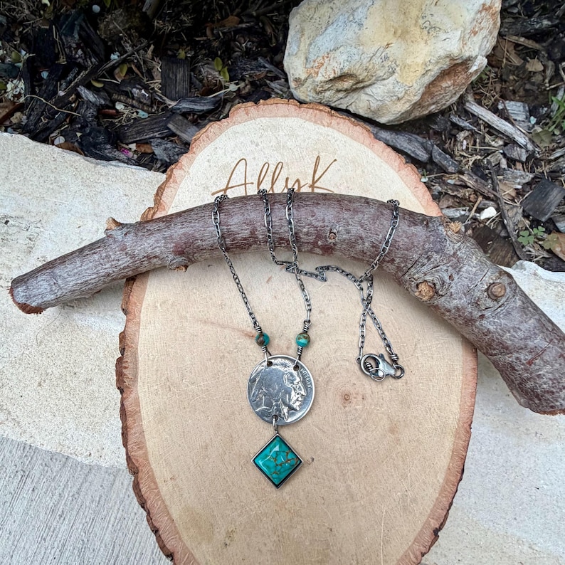 Op de afbeelding: Een zilveren ketting met een munt hanger en een turkoois edelsteen. De ketting heeft een ketting met een sluiting en wordt gepresenteerd op een houten oppervlak met de tekst "Allyk". De turkoois edelsteen heeft een vierkante vorm.