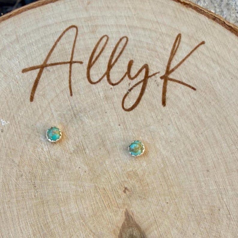 May include: A pair of small turquoise stud earrings with a silver-colored scalloped edge. The earrings are set on a light-colored wooden surface with the handwritten name "Allyk" in brown script.