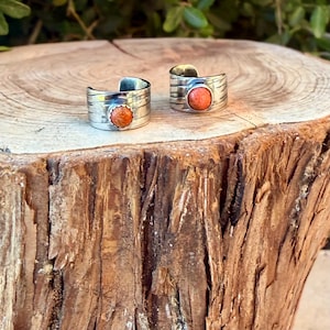 May include: Two open-ended silver rings featuring a wide, textured band with horizontal grooves. Each ring is centered with a single, round, orange-colored stone set in a scalloped bezel, creating a cohesive look for these two pieces of jewelry.