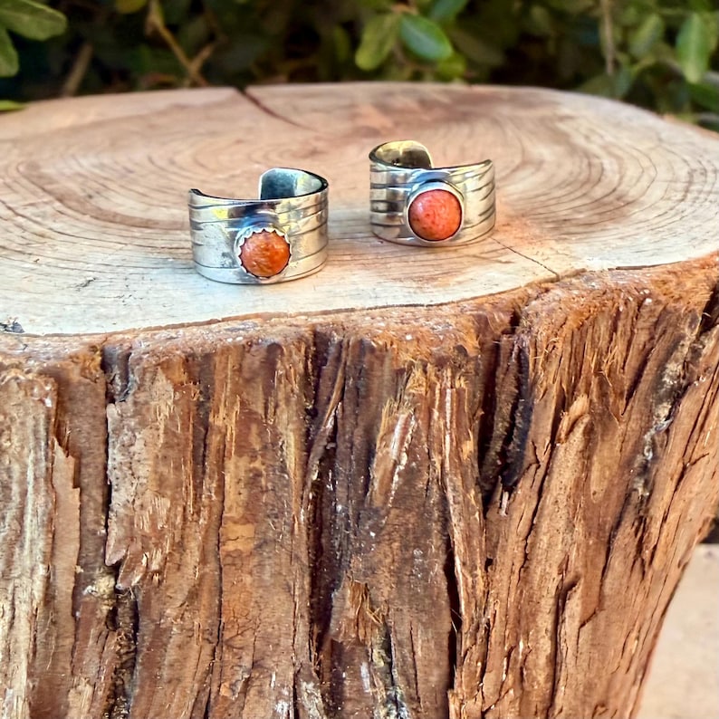 May include: Two open-ended silver rings featuring a wide, textured band with horizontal grooves. Each ring is centered with a single, round, orange-colored stone set in a scalloped bezel, creating a cohesive look for these two pieces of jewelry.