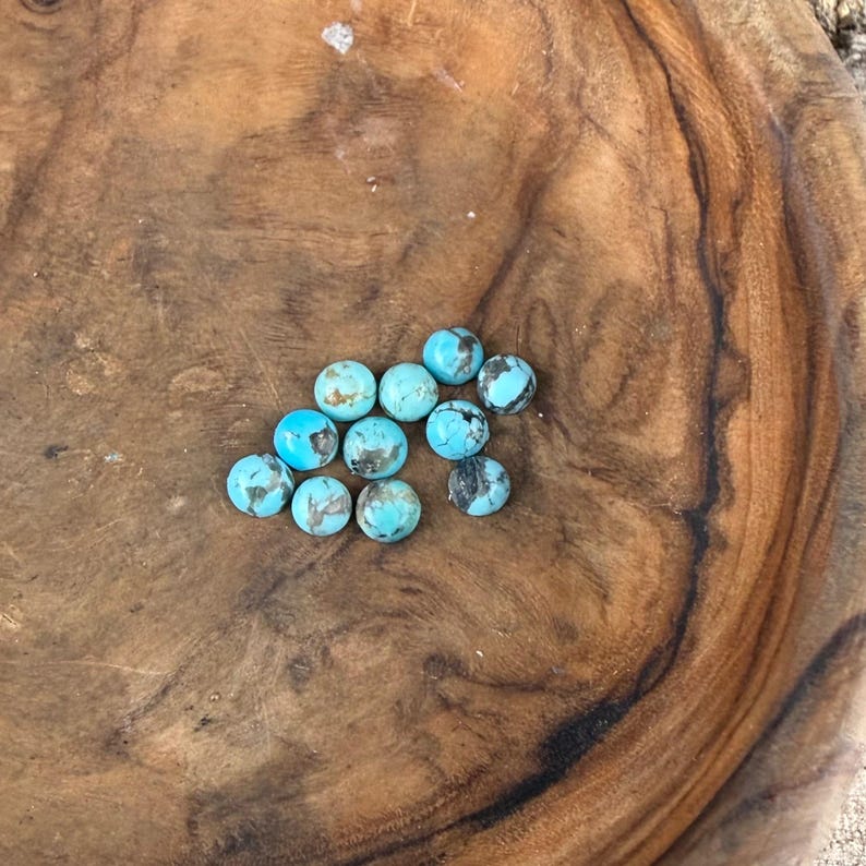 May include: A collection of round turquoise gemstones with dark brown and black matrix patterns. The stones are scattered on a dark brown wooden surface, showcasing their natural colours and textures.