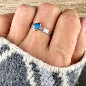 May include: A silver ring with a square, blue gemstone. The band features a floral design. The ring is worn on a finger, with a patterned gray and white sweater sleeve in the background.