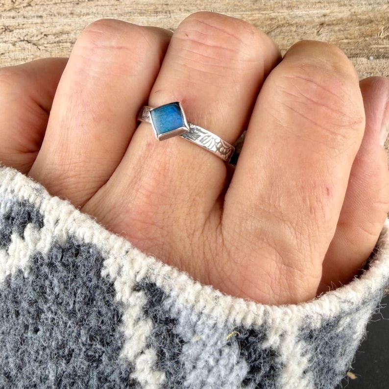May include: A silver ring with a square, blue gemstone. The band features a floral design. The ring is worn on a finger, with a patterned gray and white sweater sleeve in the background.
