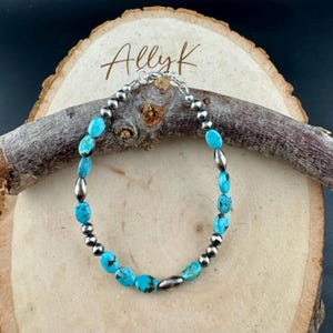 May include: Turquoise and silver beaded bracelet. The bracelet features oval turquoise stones and small silver beads, connected by a silver chain. The bracelet is displayed on a wooden surface with the text "Allyk" in cursive.