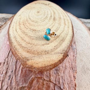 May include: A pair of gold-colored stud earrings with oval-shaped, turquoise-colored stones. The earrings are set on a wooden surface with a natural wood grain pattern. The earrings are small and delicate, with a simple design.
