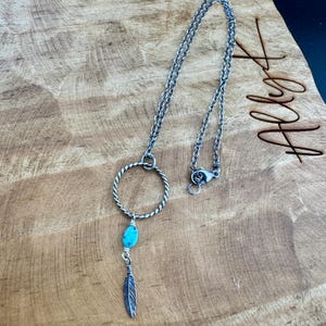 May include: A silver-toned necklace with a turquoise stone and feather pendant. The necklace features a circular design and a chain with a clasp. The pendant is suspended from the circle and is set against a wooden background.