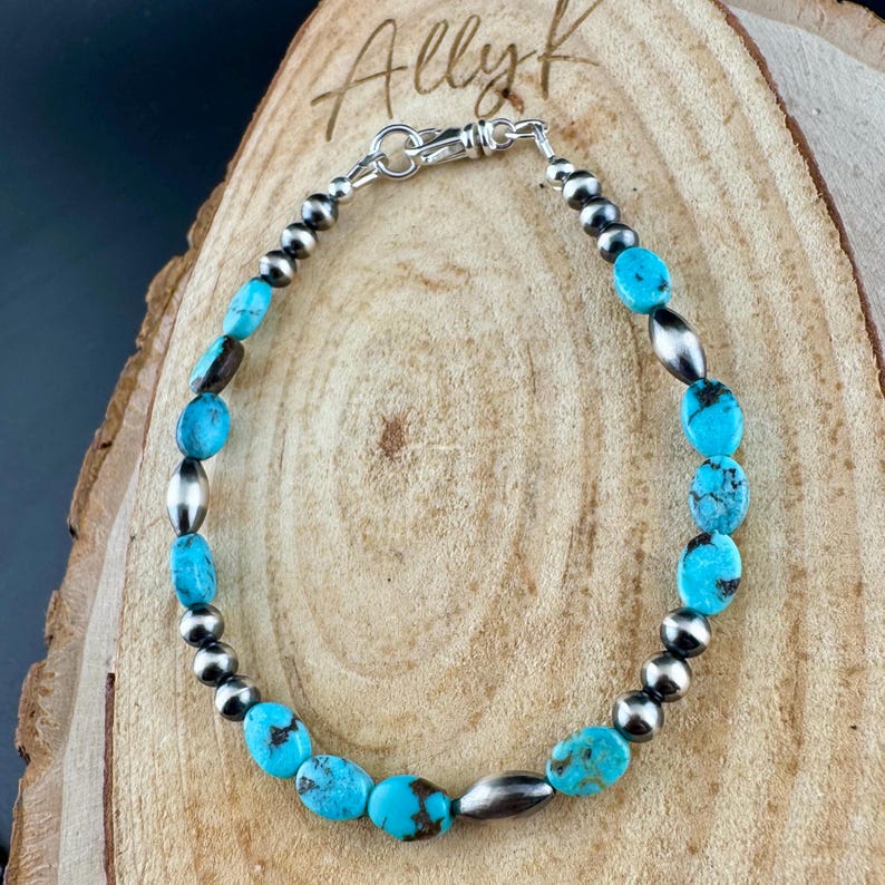 May include: A turquoise and silver beaded bracelet. The bracelet features oval turquoise stones interspersed with silver beads and elongated silver beads. The clasp is silver. The bracelet is displayed on a wooden surface. The text "Ally K" is in the upper left corner.