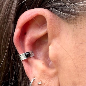 May include: Silver ear cuff with a black gemstone. The cuff features an engraved design and is worn on the ear. The image shows a close-up of the ear and the jewelry.