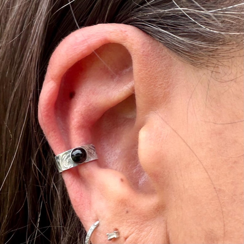 May include: Silver ear cuff with a black gemstone. The cuff features an engraved design and is worn on the ear. The image shows a close-up of the ear and the jewelry.