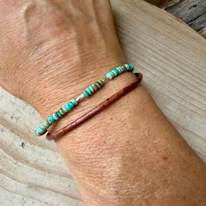 May include: A layered bracelet set consisting of one strand of small, round, turquoise-colored beads with brown matrix inclusions and one plain, smooth, dark brown leather cord strand, both worn together on the wrist as a stacked accessory.