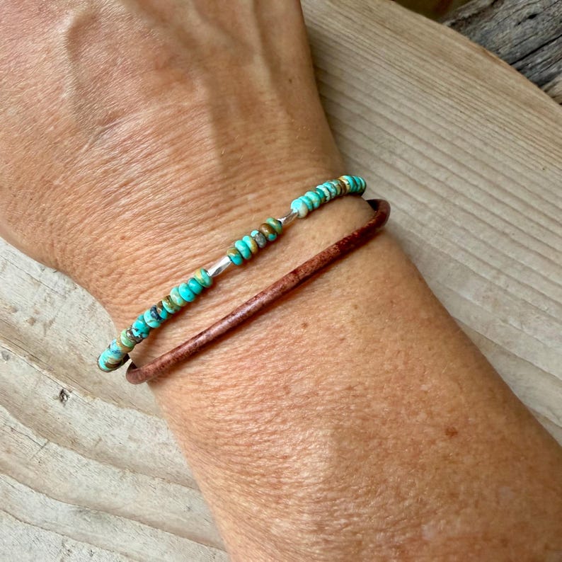 May include: A layered bracelet set consisting of one strand of small, round, turquoise-colored beads with brown matrix inclusions and one plain, smooth, dark brown leather cord strand, both worn together on the wrist as a stacked accessory.