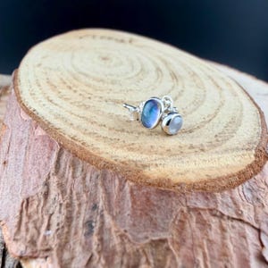 May include: A pair of silver stud earrings featuring two moonstone gemstones. One is an oval shape with blue and purple hues, and the other is a smaller, round, white stone. The earrings are displayed on a wooden surface.