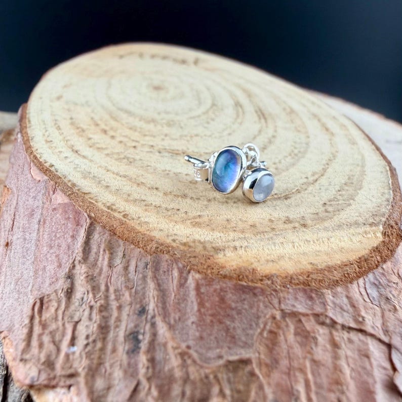 May include: A pair of silver stud earrings featuring two moonstone gemstones. One is an oval shape with blue and purple hues, and the other is a smaller, round, white stone. The earrings are displayed on a wooden surface.