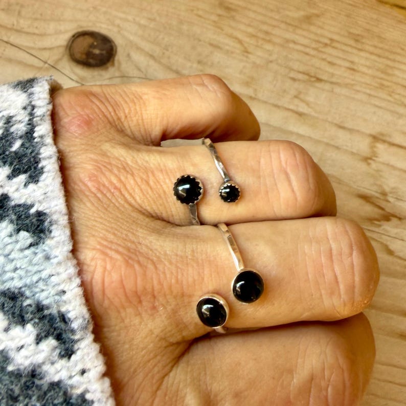 May include: Two open-style silver rings, each featuring two round black stones set at the ends of the band. The rings are worn on adjacent fingers, showcasing a polished metallic finish and smooth, dark circular gemstones in a simple, open design.