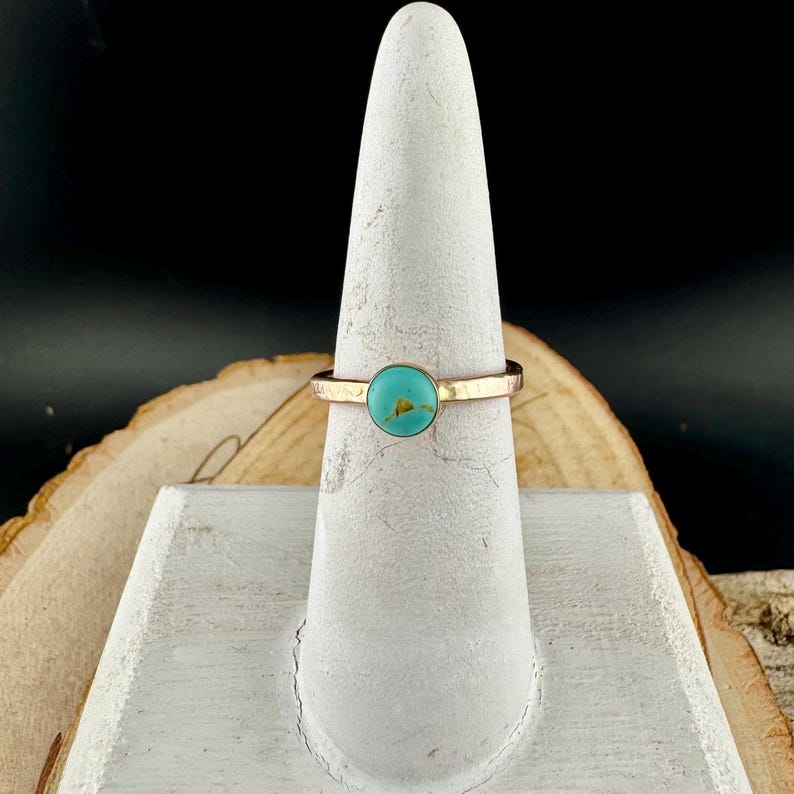 May include: A gold ring featuring a circular turquoise stone. The ring is presented on a white display stand. The band has a textured finish, and the bright turquoise stone is the focal point. The ring is displayed against a dark background.