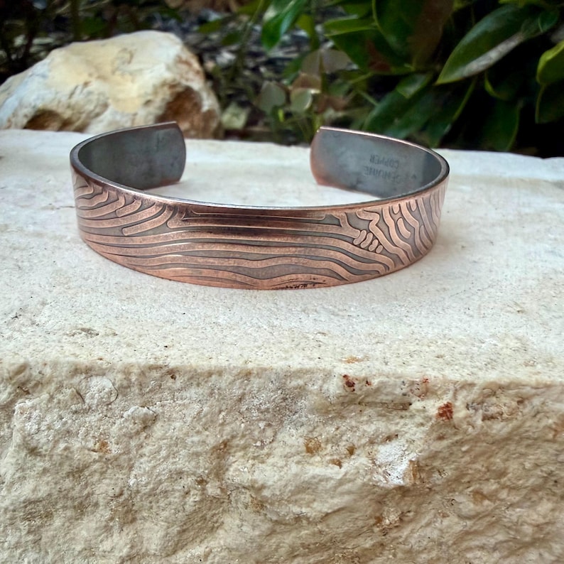 May include: Copper cuff bracelet with a textured, wave-like pattern. The open-ended bracelet has a brushed metal interior and a polished exterior. The design features a series of horizontal lines and a stylized design on the front. The bracelet is displayed on a stone surface.