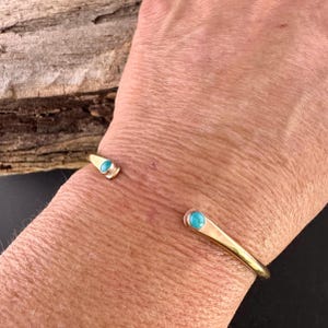 May include: A gold-colored cuff bracelet with turquoise-colored stones at each end. The bracelet has a simple, elegant design. The turquoise stones are small and round, adding a pop of color to the gold. The bracelet is shown on a person's wrist.